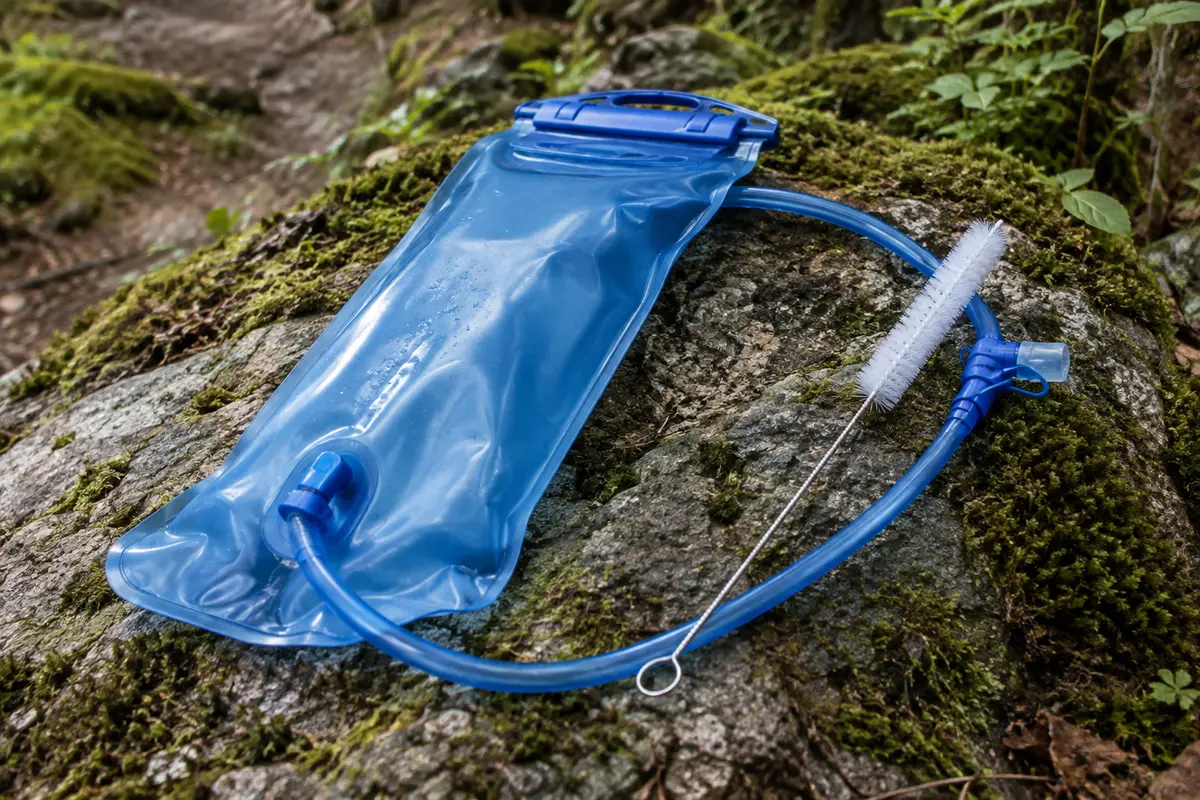 Poche d’hydratation bleue de 3 L avec tuyau, embout et brosse, posée sur une pierre près d’un sentier forestier.