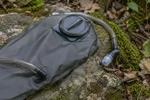 Poche à eau gris carbone de 2 litres avec tuyau flexible et valve, posée sur une pierre en forêt.