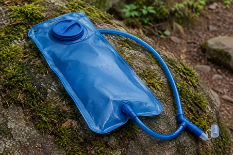Poche à eau bleue de 2 L avec tuyau et valve à mordre, posée sur une pierre près d’un sentier forestier.