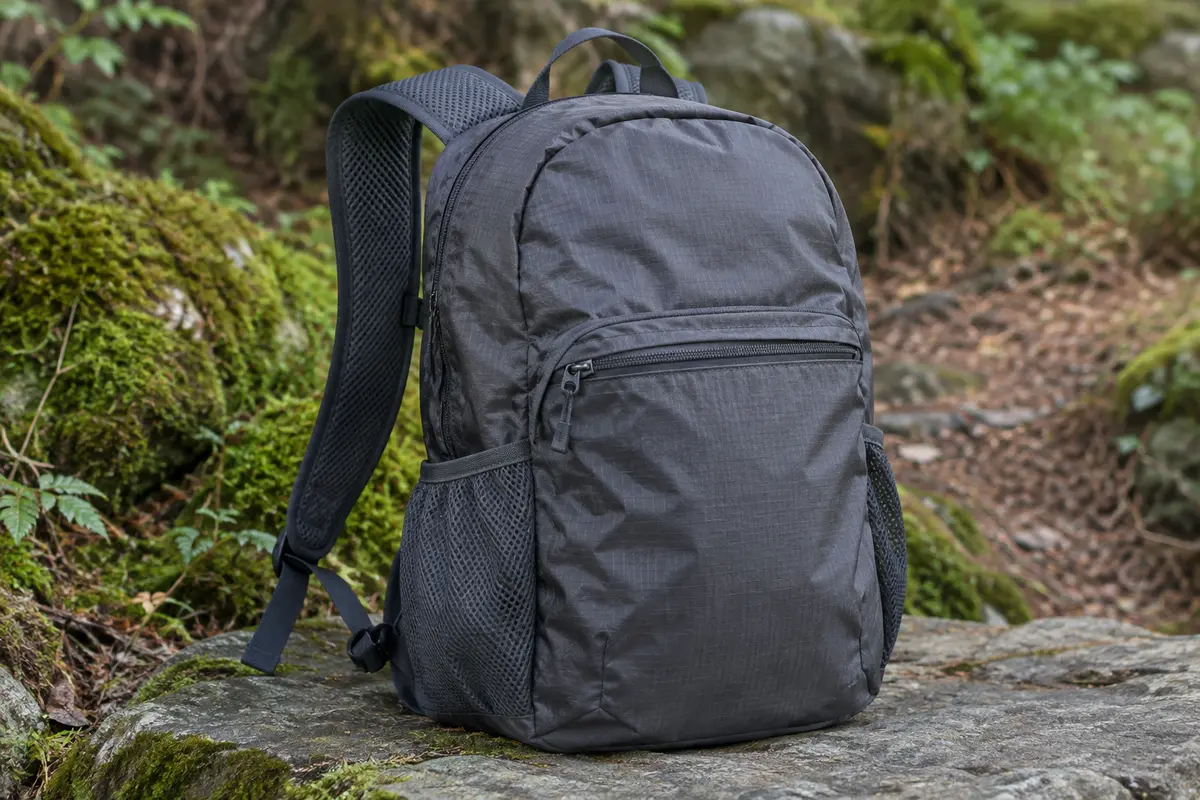Sac à dos pliable gris foncé posé sur une pierre au bord d’un sentier forestier
