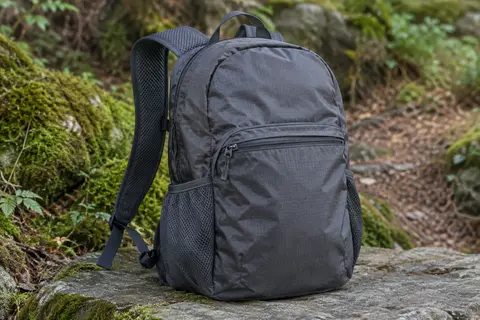 Sac à dos pliable gris foncé posé sur une pierre au bord d’un sentier forestier