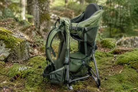 Porte-enfant de randonnée vert posé seul sur un sentier forestier, avec son cadre pliable visible.